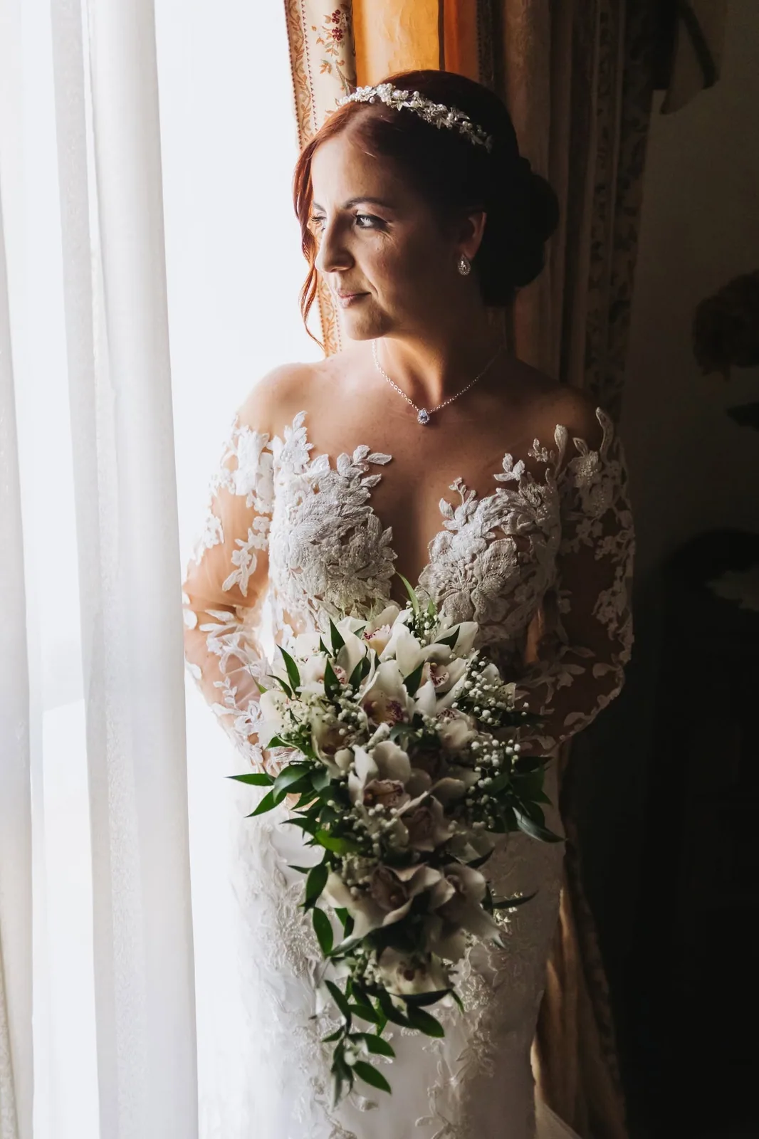 A bride in a lace wedding dress holding her bouquet beside a softly lit window in Malta
